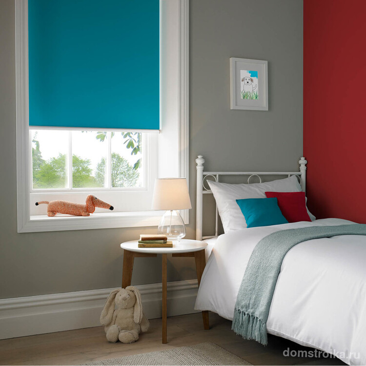 Красный цвет акцентной стены в детской комнате дополняет голубая роллета и декоративные подушки