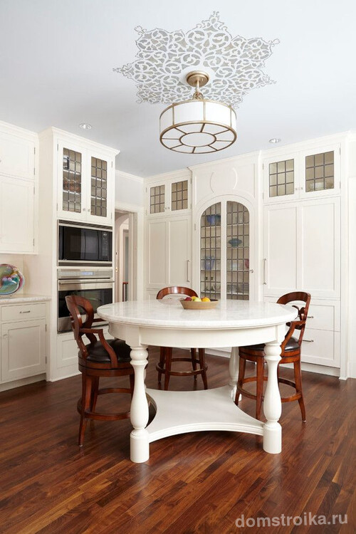 Кухня с однотонным белым гарнитуром. Белый круглый деревянный стол и коричневые деревянные высокие стулья с черными круглыми сиденьями – одинаковая форма и удачное цветовое сочетание