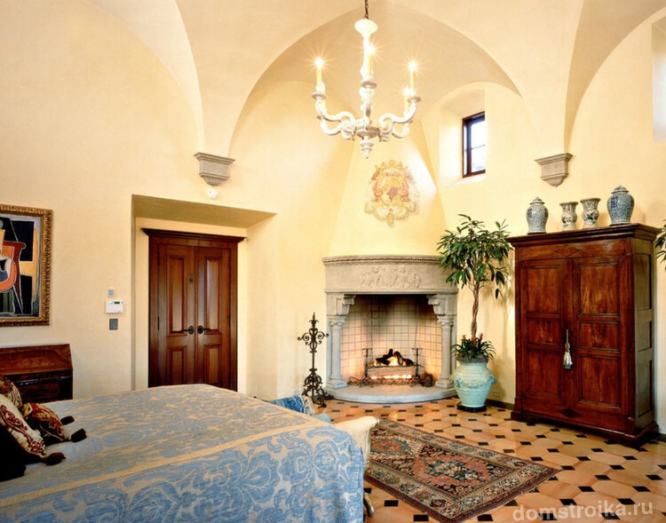Нестандартный подход к оформлению спальни в готическом стиле: кованный подсвечник, рисунок герба над камином, а также светлая отделка стен и современные растения в комнате