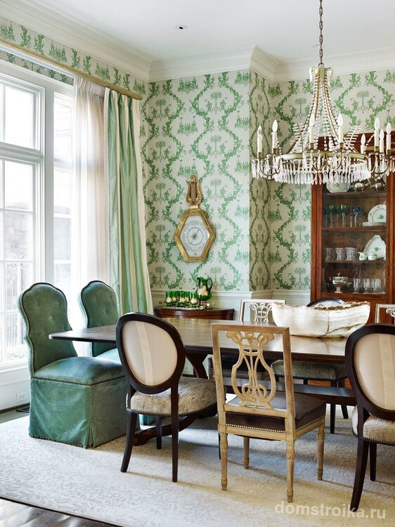 Классическая столовая комната с деревянным сервантом для посуды, хрустальной люстрой, стульями разных форм и текстильных покрытий, белыми обоями с зеленым орнаментом