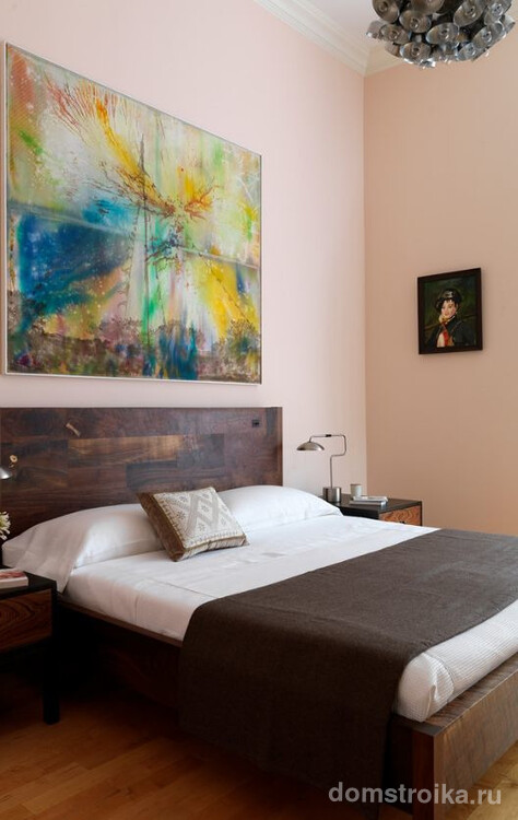 Картина с яркой абстракцией гармонично сочетается с деревянным изголовьем кровати и пастельным цветом стены